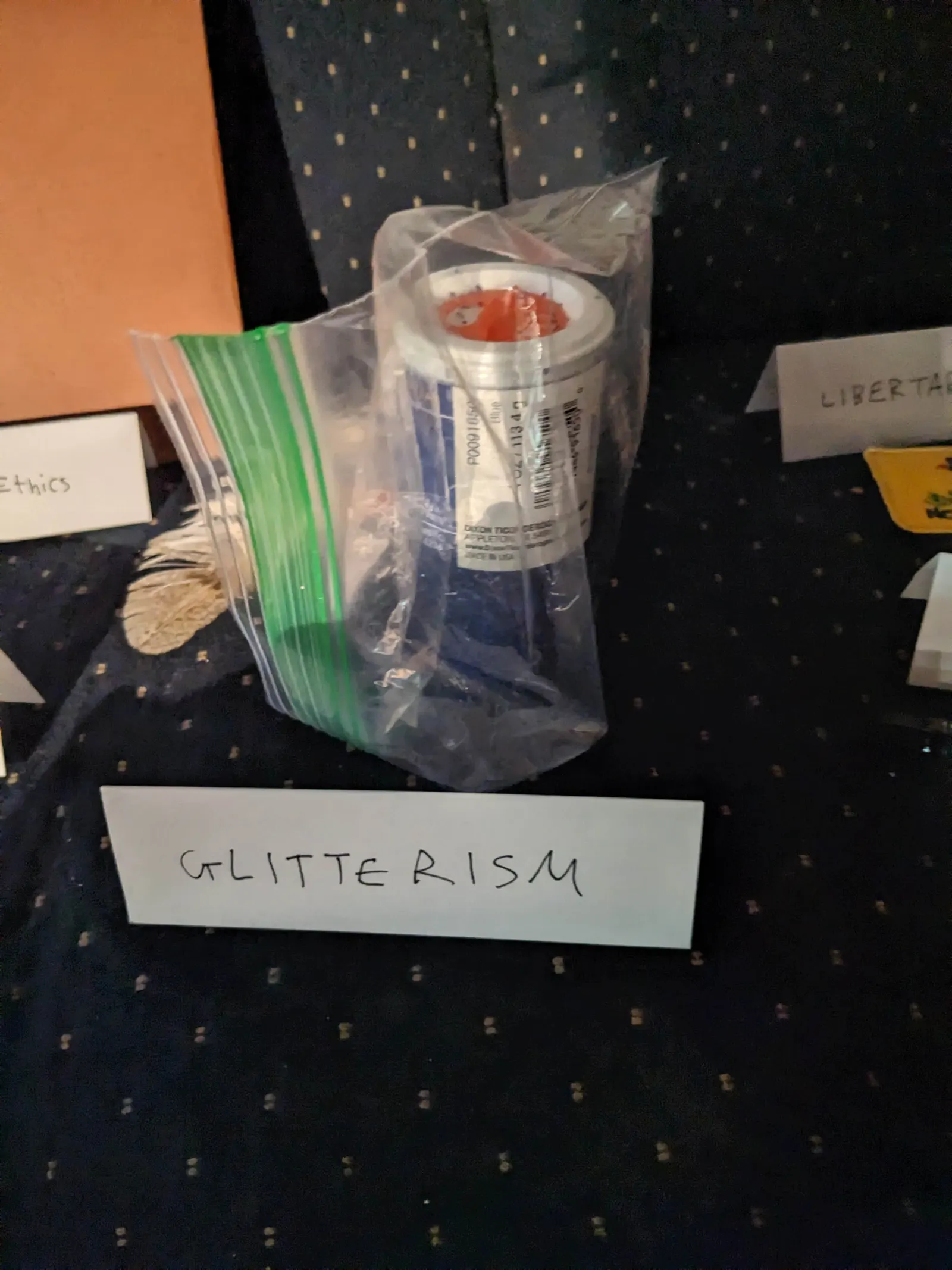 A bagged glitter object on the Ideology Altar with a tent sign labeled Glitterism.