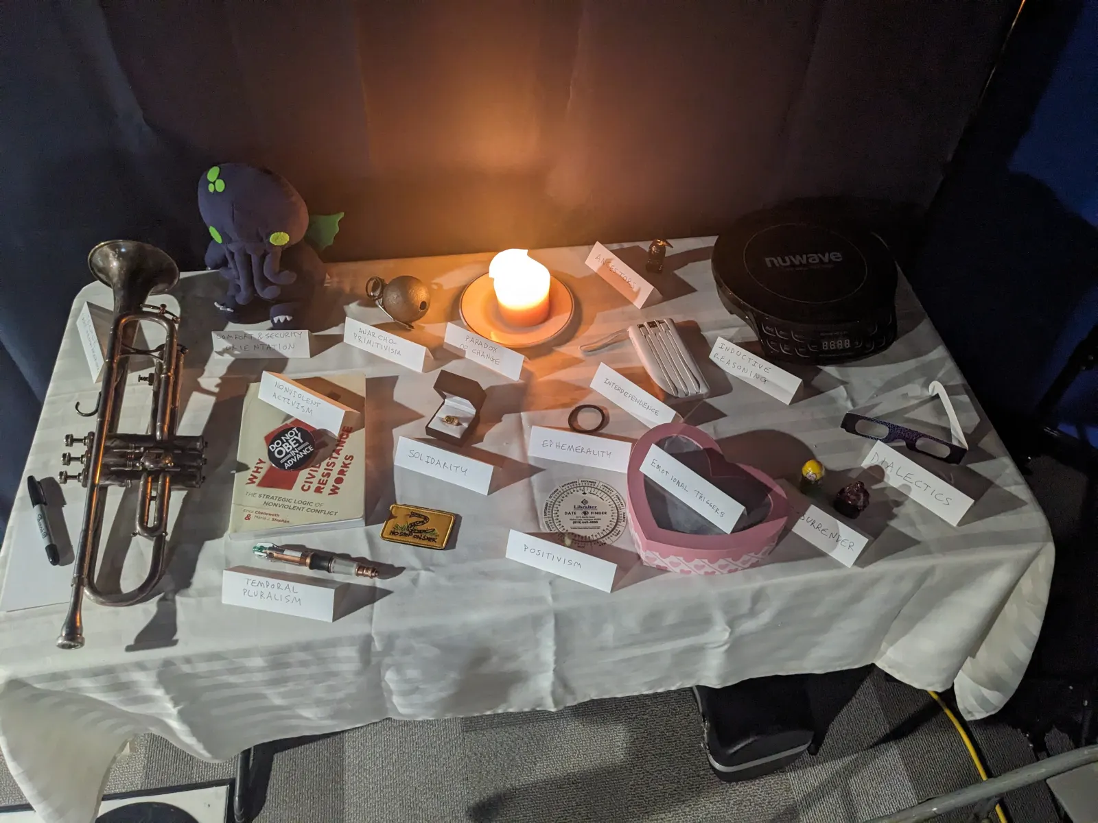 The 2025 Ideology Altar, with many participant-contributed objects and handwritten tent signs arranged together on a long table.