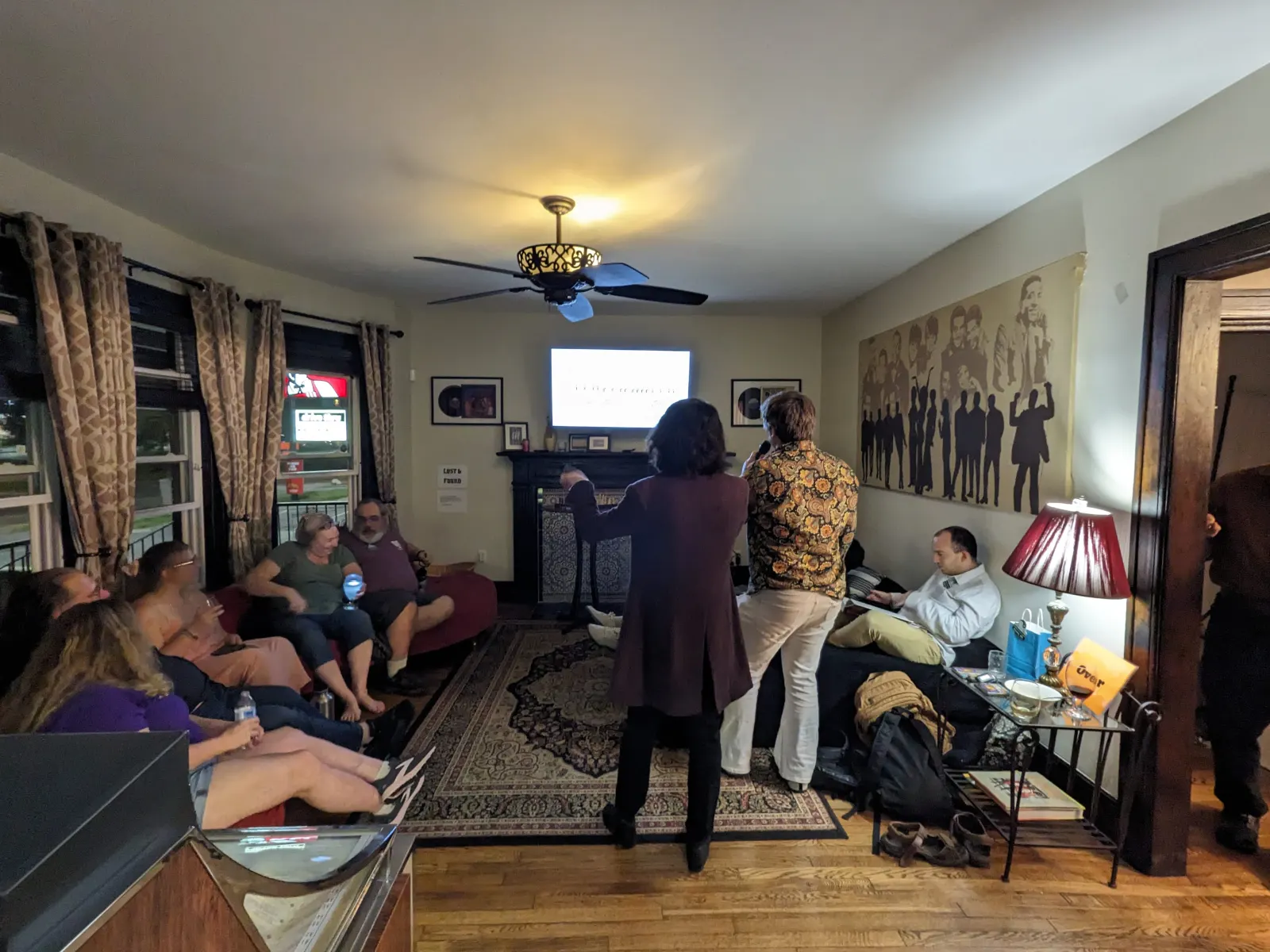 Attendees seated around a living room while two people present beside a screen.